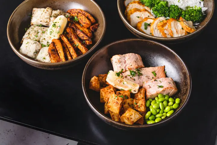 Four bowls of cooked food on a dark table, each with fish or meat, a grain and something green.