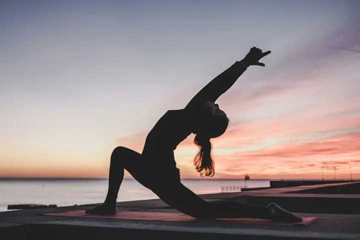 A woman in silhouette holding a low lunge on a mat, against a pink sunset over water.