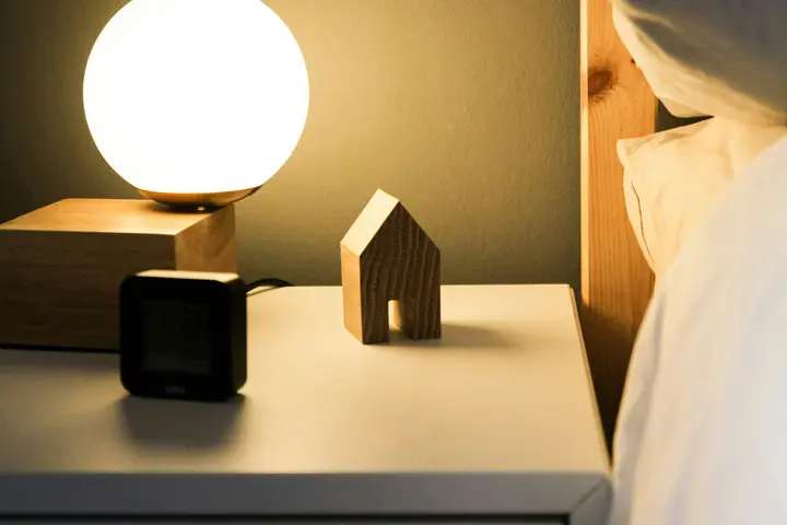A bedside table at night: a warm round lamp, a small clock, and the corner of a bed.